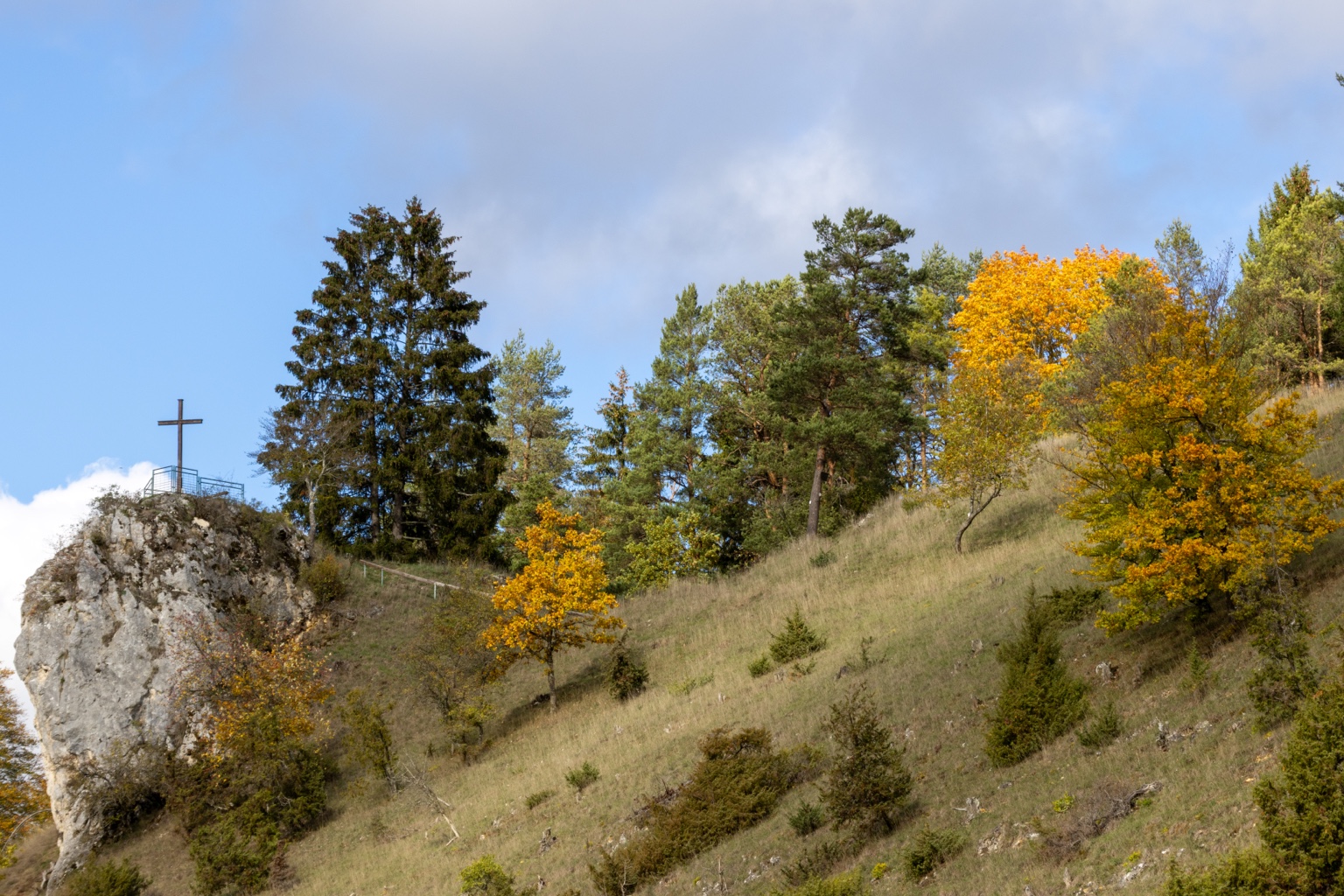 Heller Felsen mit einem Kreuz neben einem Hang mit goldgelben Laubbäumen und grünen Nadelbäumen