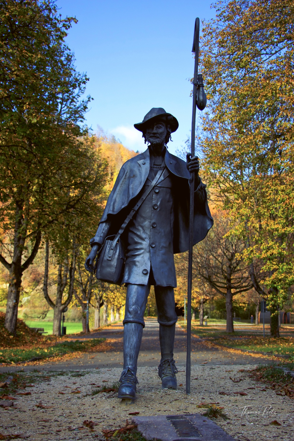 Figur mit Hut, Tasche und langem Stab vor herbstlichen Bäumen