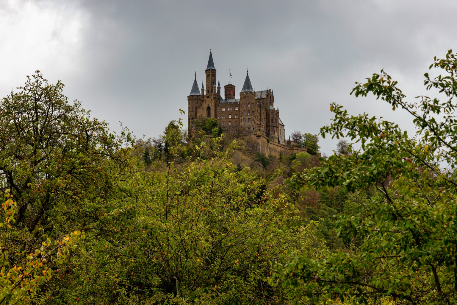 Die Mauern und spitzen Türme der Burg Hohenzollern über grünen und gelben Obstbäumen vor einem grauen Himmel