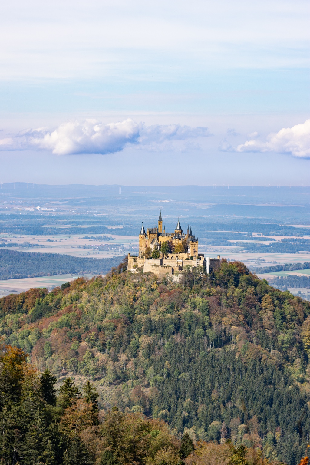 Burg Hohenzollern mit Türmen und Mauern auf einem herbstlich bewaldeten Berg vor Feldern, Wäldern und Wolken