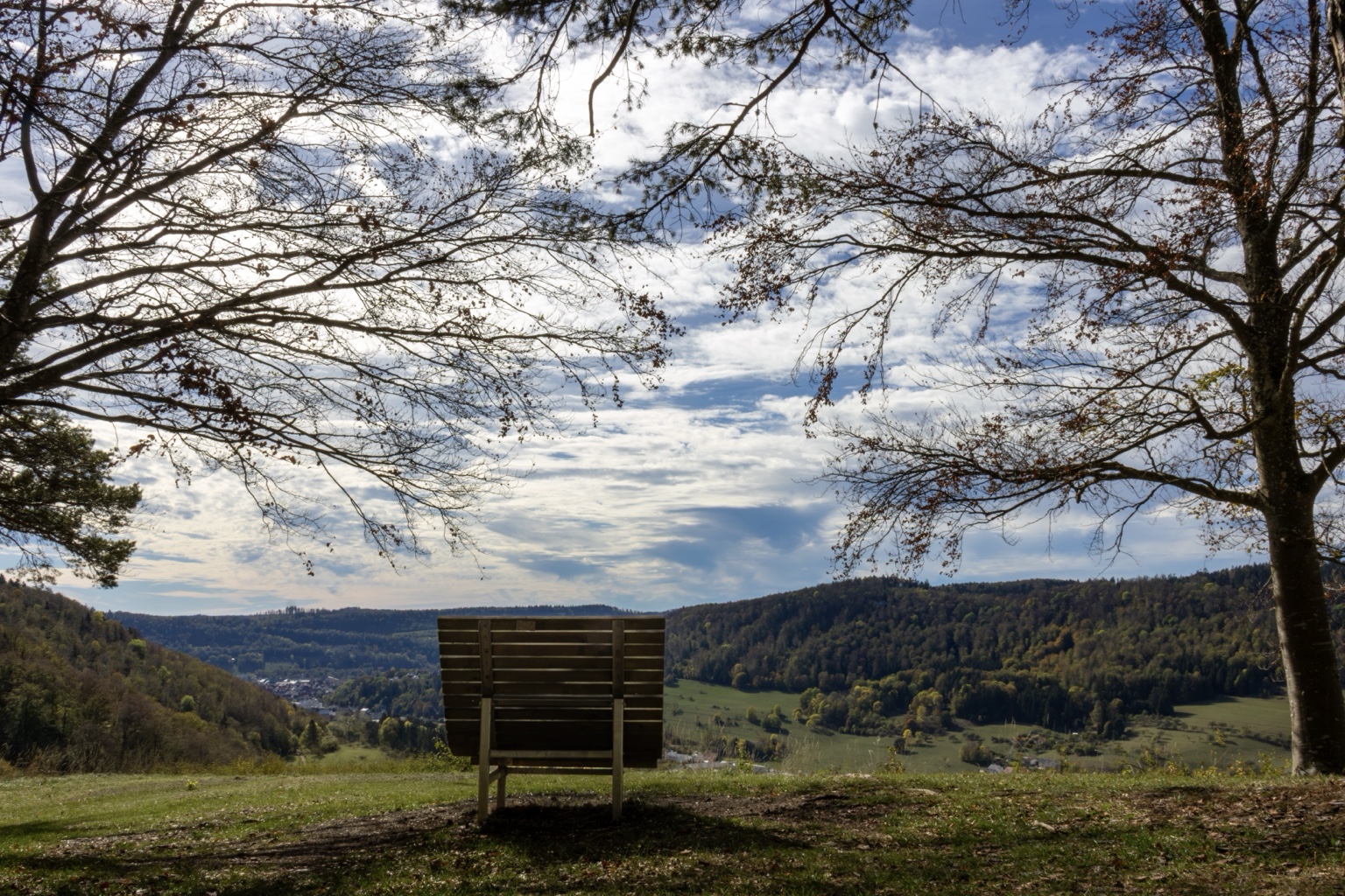 Holzliege zwischen Bäumen mit Aussicht auf Wiesen, Häuser und bewaldete Hügel