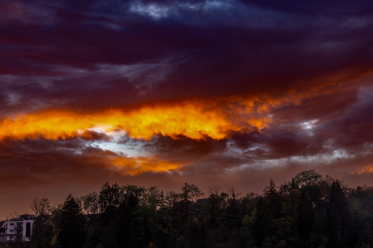 Breites orangegoldenes Wolkenband zwischen dunklen violetten Wolken über einem bewaldeten Hang