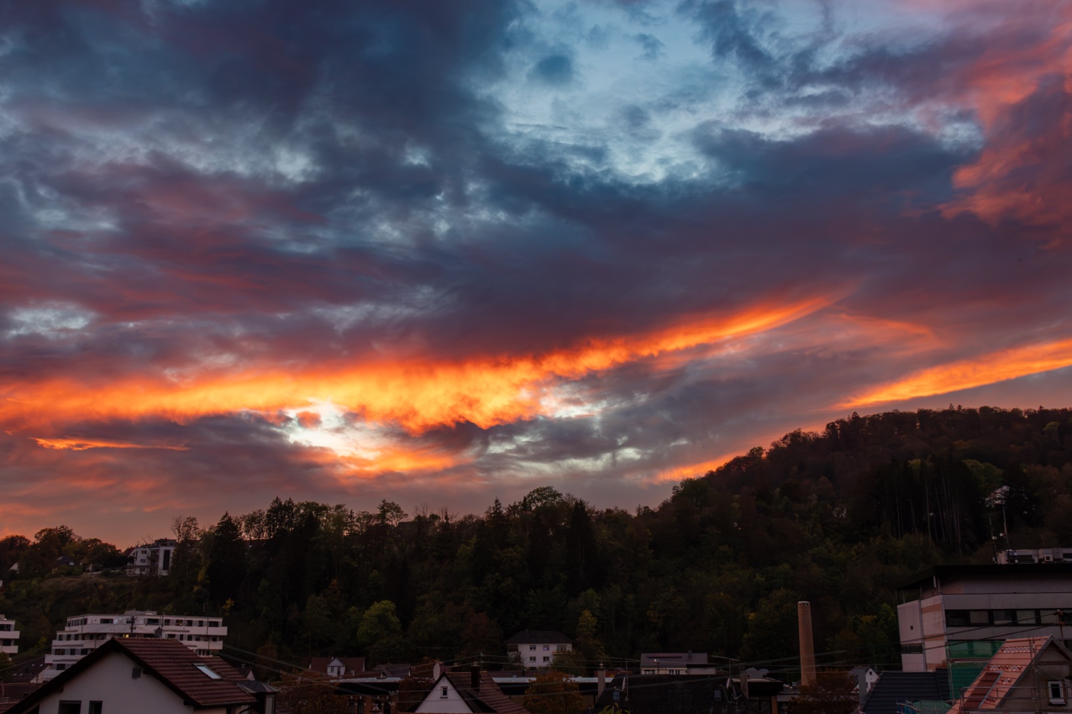Weiter Abendhimmel mit orange leuchtendem Wolkenband über Häuserdächern und einem bewaldeten Hügel