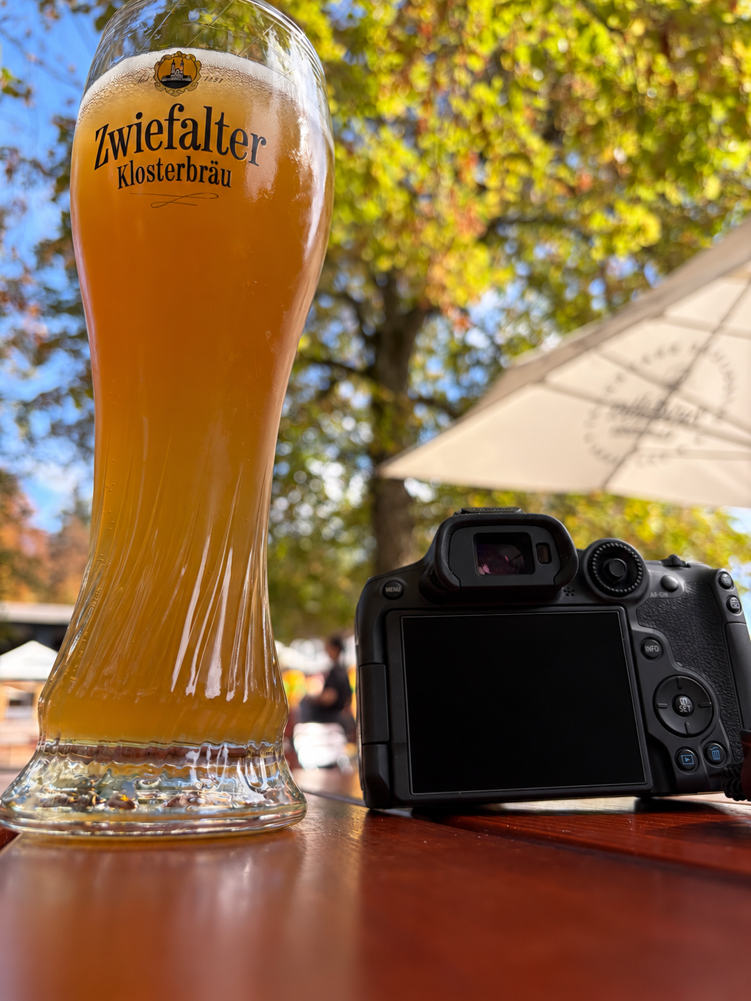 Glas mit goldenem Bier neben einer Kamera mit verdecktem Display auf einem Holztisch unter herbstlichen Bäumen
